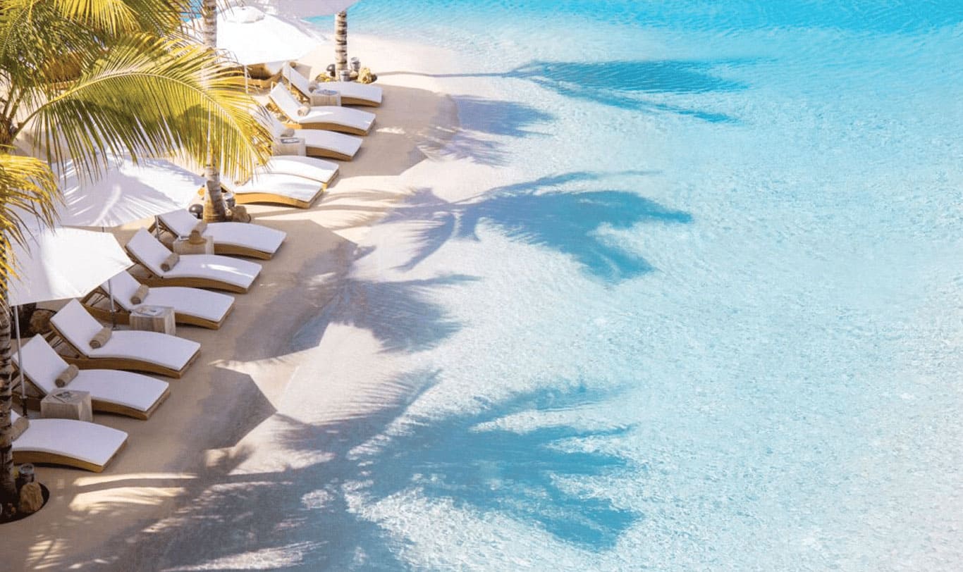 Piscine d'hotel en quartz blanc se jetant dans la mer des caraibes. Chaises longues, parasols et palmiers