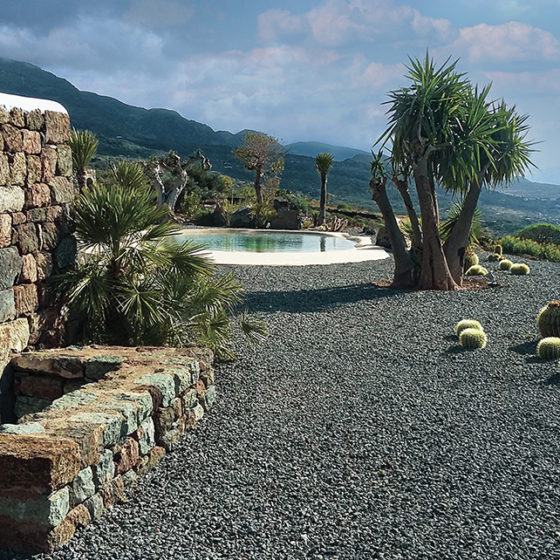 piscine lagon derrière une maison traditionnelle en pierre sur lit de pouzzonale et vue sur la mer avec palmiers et cactus