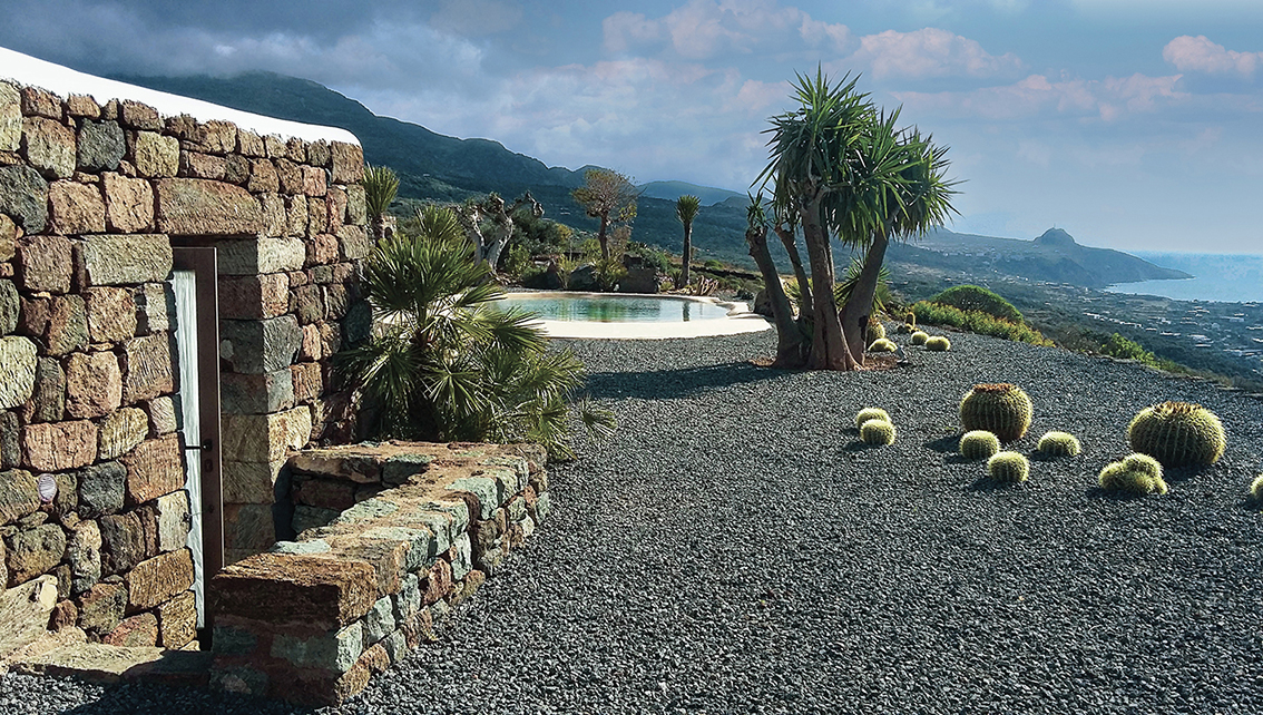 piscine lagon derrière une maison traditionnelle en pierre sur lit de pouzzonale et vue sur la mer avec palmiers et cactus